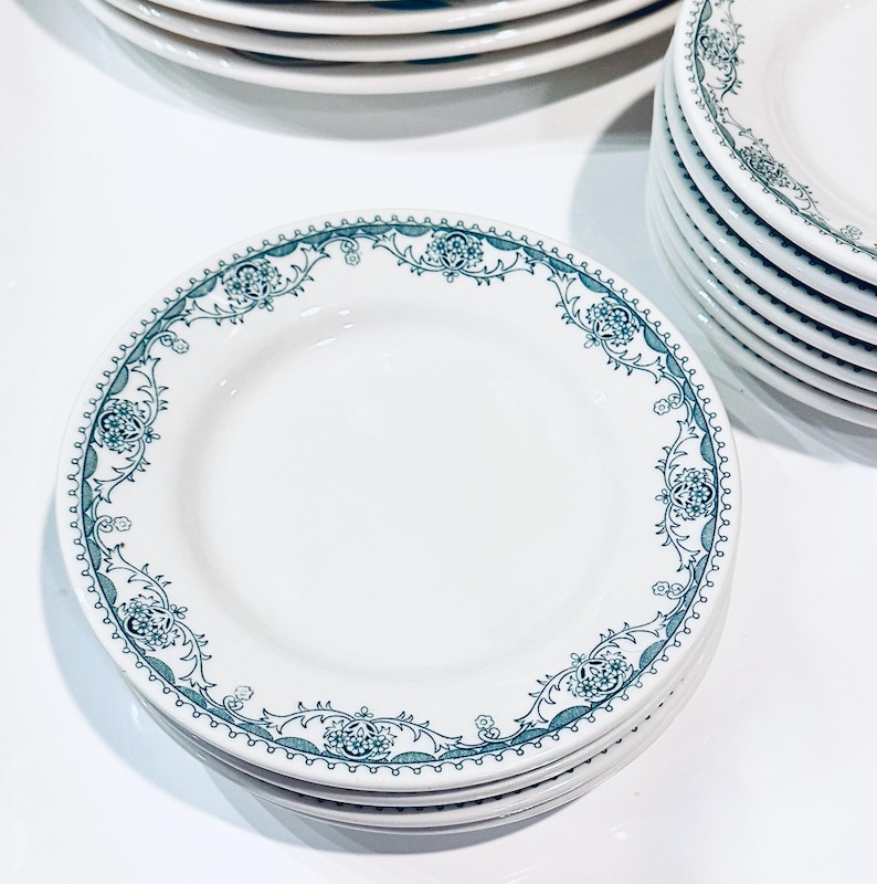 Może przedstawiać: Stos białych ceramicznych talerzy z zielonym wzorem kwiatowym wok&oacute;ł krawędzi.