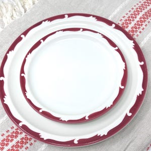 Op de afbeelding: Twee witte keramische borden met een bordeauxrode rand en een decoratief golfpatroon. Het kleinere bord is genest in het grotere bord.