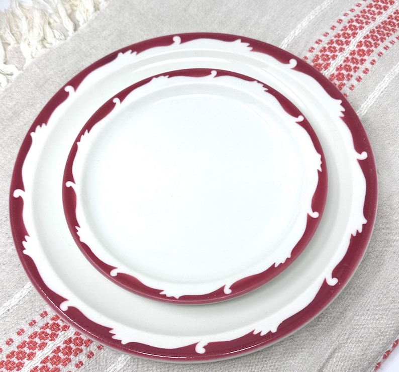 Op de afbeelding: Twee witte keramische borden met een bordeauxrode rand en een decoratief golfpatroon. Het kleinere bord is genest in het grotere bord.