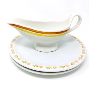 May include: A white ceramic gravy boat with a handle, featuring brown and yellow stripes near the rim. It sits on a white plate with a floral pattern in orange. The set is designed for serving sauces and gravies.