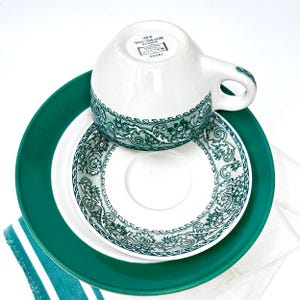 May include: An ivory-colored teacup, saucer, and plate set with a green floral design. The teacup is upside down, resting on the saucer, which is on top of a larger green plate. The set is on a white background with a teal and white striped cloth.