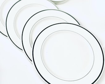 May include: A set of white ceramic plates arranged in a staggered, overlapping row. Each circular plate features a smooth, glossy finish and a single, thin, dark navy blue band encircling the outer rim, creating a clean and uniform appearance.
