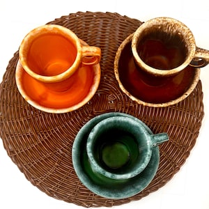 May include: Three ceramic cups and saucers in orange, brown, and dark green, each featuring a glossy glaze with a lighter, mottled rim. The cups have side handles and are arranged in a triangular formation, showcasing a smooth, reflective finish.