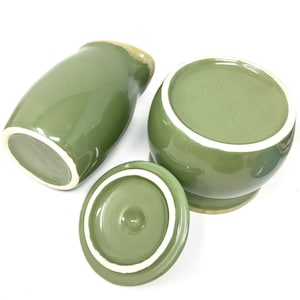 May include: A set of three green ceramic kitchenware items. The set includes a lidded jar, a small round jar, and a cylindrical container. The items have a glossy finish with a white interior and a light brown rim.