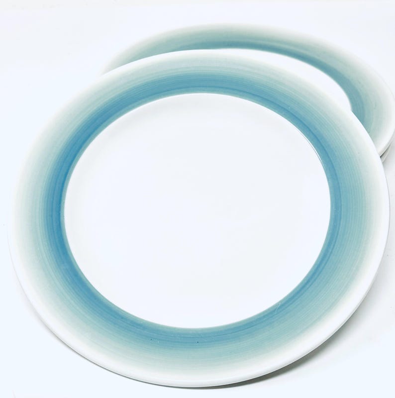 May include: Two white ceramic plates with a gradient blue border. The blue color transitions from a darker shade at the edge to a lighter shade towards the center. The plates are set against a white background.