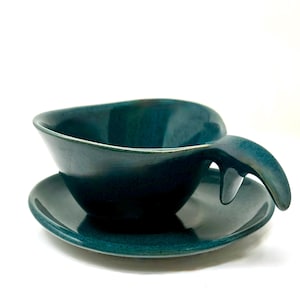 May include: A deep teal ceramic cup and saucer set with a glossy finish. The cup features an irregular, organic rim shape and a thick, curved handle that extends outward from the side. The saucer is round with a matching deep teal glaze.
