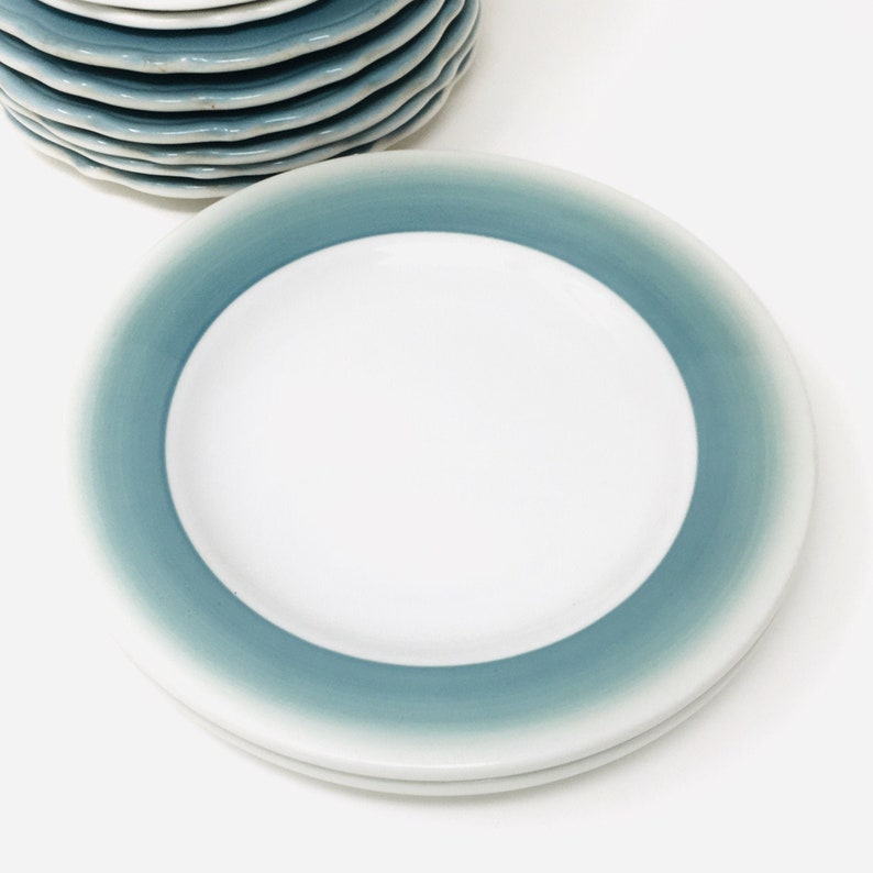 May include: A stack of white ceramic plates with a teal blue gradient border. The plates are round with a slightly wavy edge. The plates are stacked on a white surface.