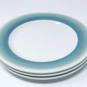 May include: A stack of four white ceramic plates with a blue and teal gradient border. The plates are round and have a simple, modern design. The gradient color transitions from a light teal to a darker blue.