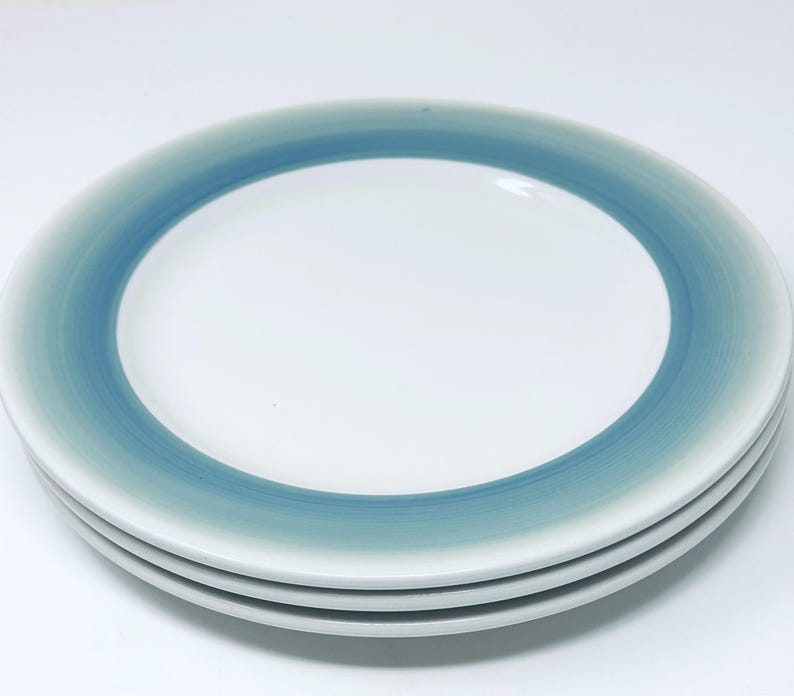 May include: A stack of four white ceramic plates with a blue and teal gradient border. The plates are round and have a simple, modern design. The gradient color transitions from a light teal to a darker blue.