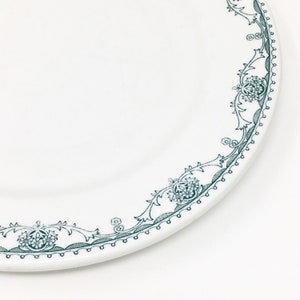 Może przedstawiać: Biały talerz ceramiczny z zielonym wzorem kwiatowym na brzegu. Talerz ma falisty brzeg i podwyższony rant.