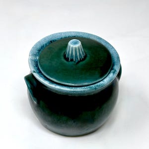 May include: A dark green ceramic jar with a matching lid featuring a light blue glazed rim and a central fluted knob. The jar has a rounded body and small side handles, showcasing a glossy finish with deep emerald and teal color variations.