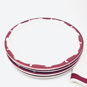 May include: Three white ceramic dinner plates with a burgundy rim and a white floral design. The plates are stacked on top of each other.