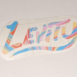 May include: A vinyl decal featuring the word Levity in a cursive script font. The lettering has a swirling, marbled pattern of pastel pink, blue, yellow, and orange colors with a subtle glitter finish. The decal is on a clear transfer sheet.