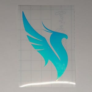 May include: A cyan iridescent vinyl decal featuring a stylized bird silhouette with extended wings, presented on a clear transfer sheet with a white grid backing. The design includes a small, faint, cursive text element visible on the right side.