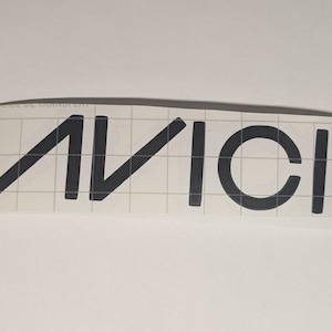 May include: Black vinyl decal featuring the word "AVICII" in a bold, sans-serif font. The letters are evenly spaced and appear on a clear backing with a grid pattern. The decal is designed for application on various surfaces.