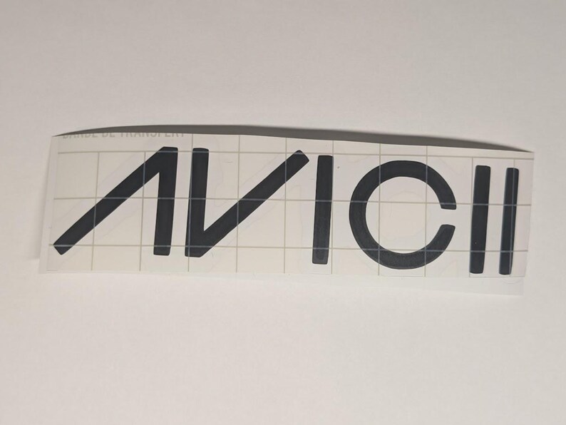 May include: Black vinyl decal featuring the word "AVICII" in a bold, sans-serif font. The letters are evenly spaced and appear on a clear backing with a grid pattern. The decal is designed for application on various surfaces.