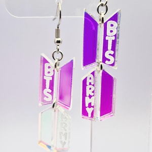Puede incluir: Un par de pendientes colgantes con el logotipo de BTS y la palabra ARMY. Los pendientes tienen un esquema de color púrpura e iridiscente con alambres para las orejas plateados. Los pendientes están hechos de un material reflectante.