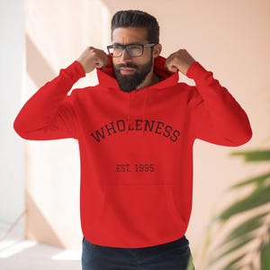 Puede incluir: Una sudadera con capucha roja brillante con la palabra "WHOLENESS" impresa en negro en el pecho. Debajo, el texto dice "EST. 1995". La sudadera tiene capucha y bolsillo delantero.