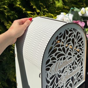 May include: A white arched wooden box featuring a laser-cut teardrop pattern on the front panel. The front displays the engraved text Matthew and Amanda in a cursive script. The top surface has a series of horizontal lines and a slot.