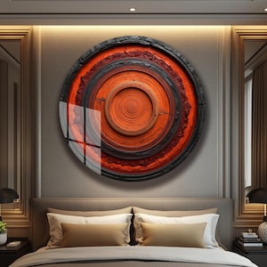 May include: A large, circular wall art piece with a layered design. The artwork features concentric circles in shades of orange, red, and black, with a textured, wood-like appearance. The piece is mounted on a neutral-colored wall above a bed.