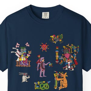 May include: Navy blue t-shirt with a vibrant graphic depicting Aztec-inspired figures. The design incorporates various characters and symbols in red, yellow, green, and purple. The shirt has a classic crew neck.