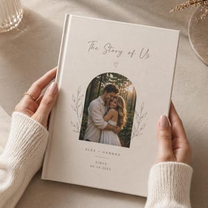 Puede incluir: Un álbum de fotos color crema titulado "The Story of Us" con una foto de una pareja abrazándose. La portada incluye los nombres "Alex + Hannah" y la fecha "05.14.2022". Elementos florales decorativos enmarcan la foto.