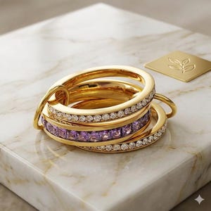 Puede incluir: Un conjunto de anillos dorados, algunos con piedras preciosas claras y moradas, entrelazados y colocados sobre una superficie de mármol. También hay un pequeño cuadrado dorado con un diseño de hojas.