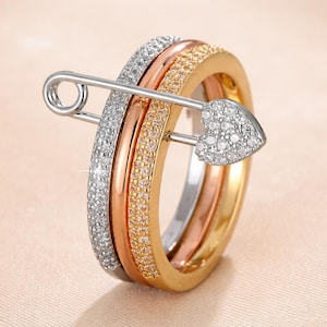 Peut inclure: Un ensemble de bagues empilables avec trois anneaux argentés, or rose et dorés, chacun incrusté de petites pierres scintillantes. Une épingle de sûreté argentée, ornée d'une tête en forme de cœur également sertie de pierres, traverse les bagues, créant un design de bijou unique.