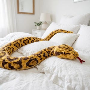Puede incluir: Un gran juguete de peluche con forma de serpiente en tonos amarillos y marrones, que se asemeja a una pit&oacute;n, descansa sobre una cama con ropa de cama blanca. El cuerpo de la serpiente est&aacute; estampado con manchas marrones oscuras, y su cabeza presenta ojos amarillos y una lengua roja.