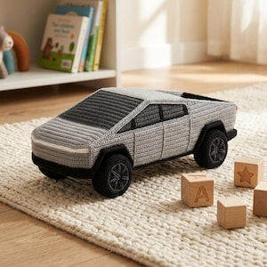 May include: A crocheted toy truck in a light gray color with dark gray accents on the roof and windshield, black wheels, and black trim. The truck features a geometric, angular body design with a flat bed and a distinct pointed front hood profile.