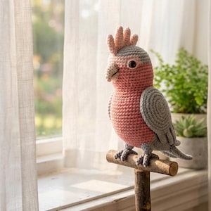 May include: A crocheted Galah cockatoo figurine featuring a soft pink chest and face, light gray wings and back, and a pale pink crest. The bird has a dark eye and a light-colored beak, and it is perched on a small, horizontal wooden branch stand.