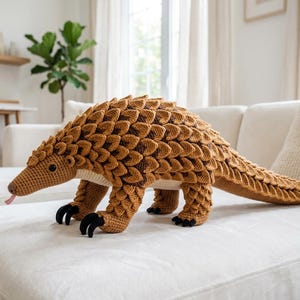 Op de afbeelding: Een grote, handgemaakte gehaakte pangolin knuffel in bruine tinten. De knuffel heeft gedetailleerde schubben, een lange staart en kleine zwarte klauwen. De pangolin heeft een lange snuit en een roze tong.