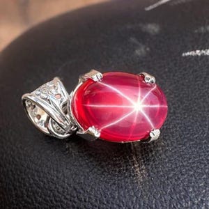 May include: A silver pendant with a large, oval star ruby. The ruby is a deep red colour, displaying a six-rayed star effect. The pendant has an ornate silver setting with intricate detailing.