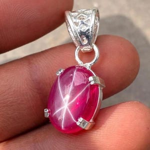 May include: An oval-shaped, vibrant pinkish-red star gemstone pendant set in a polished silver-toned metal prong setting. The cabochon stone displays a distinct six-rayed star pattern across its surface, suspended from a decorative silver bail.