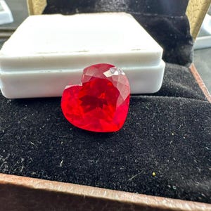 Può includere: Una gemma sfaccettata a forma di cuore di un colore rosso vibrante e traslucido. La pietra presenta una superficie lucida con molteplici tagli geometrici che riflettono la luce, creando un aspetto brillante. È esposta su una base bianca.