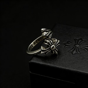 May include: A silver ring featuring a cross-shaped design and a floral accent. The ring has an open design and is set against a dark background. The ring's band features engraved text.
