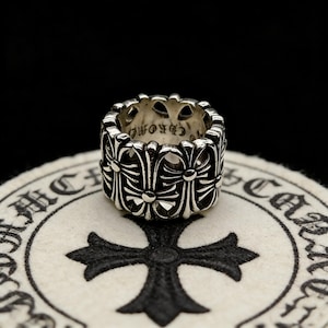 May include: A silver ring with a repeating cross design. The ring is wide and has a detailed pattern of crosses and floral elements. The ring is on a white surface with a black cross design.