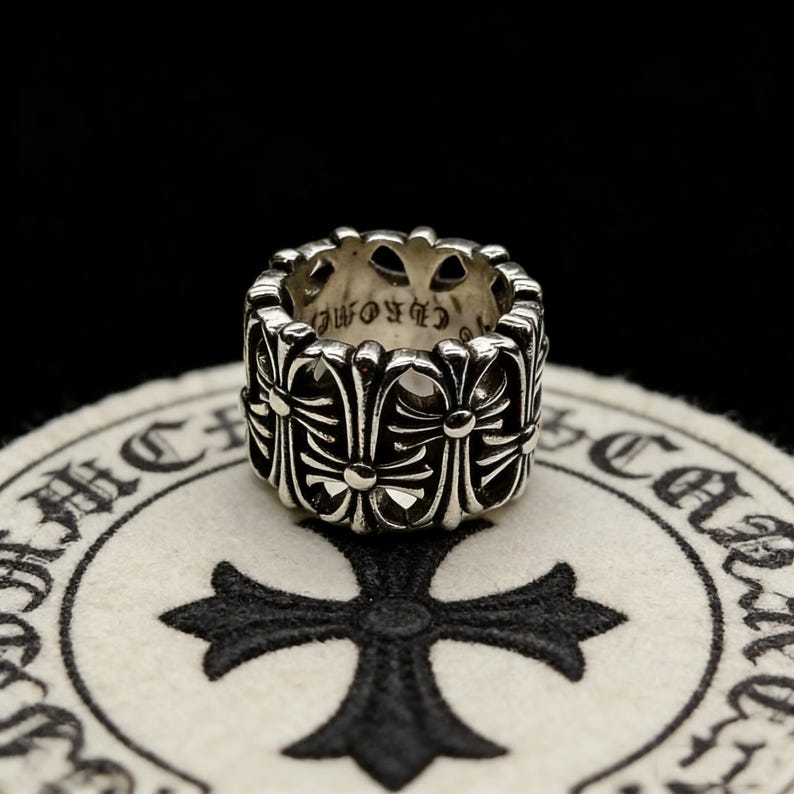 May include: A silver ring with a repeating cross design. The ring is wide and has a detailed pattern of crosses and floral elements. The ring is on a white surface with a black cross design.