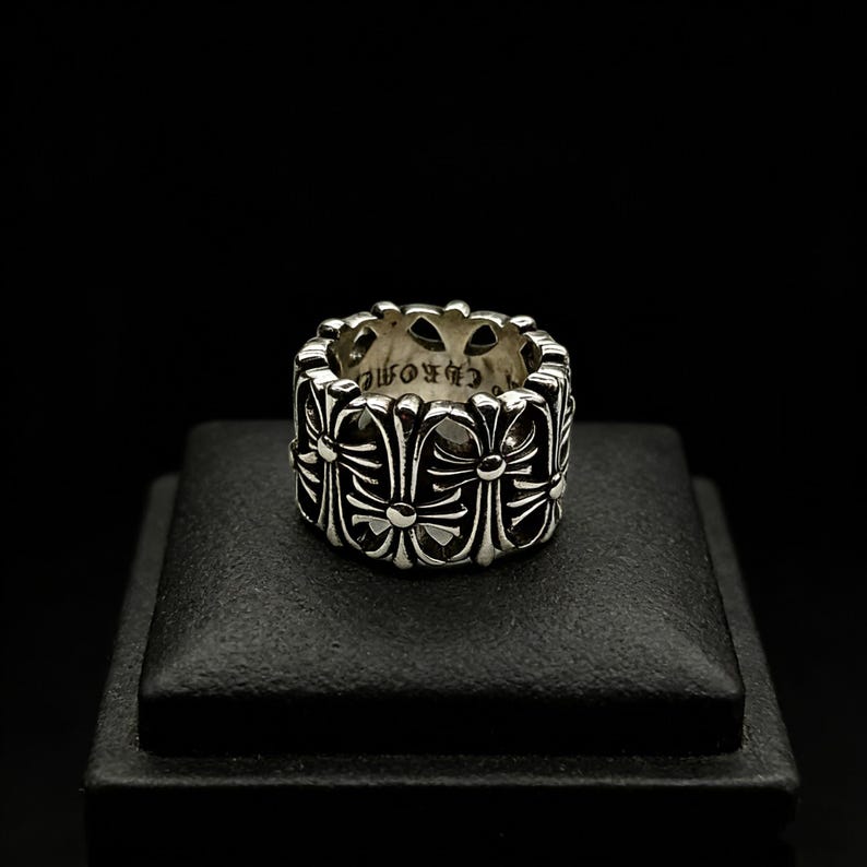 May include: A silver ring with a repeating cross design. The ring is on a black surface against a black background. The word "CHROME" is visible on the inside of the ring.