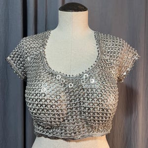 May include: A short-sleeved crop top constructed from interlocking silver-toned metal rings, creating a chainmail-like mesh pattern. The garment features a rounded neckline and a straight hem, with a consistent metallic finish throughout the piece.