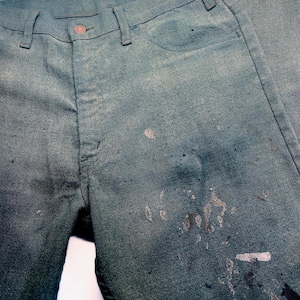 May include: Close-up of teal-colored denim jeans with paint splatters. The jeans have a button closure and classic five-pocket design. The fabric shows signs of wear and tear, with faded areas and paint marks.