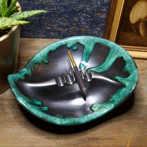 May include: A black and green ceramic ashtray with a unique design. The ashtray has a deep well and a raised rim. It is perfect for holding cigarettes or other small items.