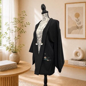 May include: A black kimono-style jacket with wide sleeves, displayed on a mannequin. The jacket features a floral embroidery detail on the front and decorative white closures. The mannequin is positioned in a well-lit room with a neutral color palette.