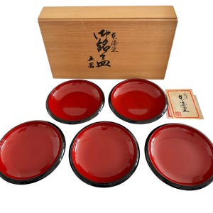 May include: A set of five round plates with a glossy red interior and a black exterior rim. The plates are arranged in front of a rectangular wooden storage box featuring black Japanese calligraphy on the lid and a small paper card with text.