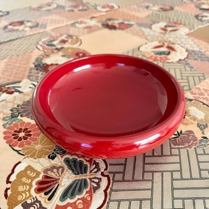 May include: A glossy, bright red serving bowl. The round bowl sits on a patterned fabric with floral and geometric designs. The bowl's smooth surface reflects light, highlighting its shape and colour. The fabric is in shades of beige and brown.