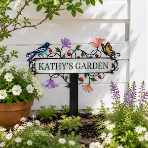 Puede incluir: Un letrero de jardín rectangular con marco de metal negro que presenta el texto KATHY'S GARDEN en letras verdes. El letrero está decorado con un pájaro azul, una mariposa amarilla y negra, flores moradas y naranjas y pequeñas bayas rojas.