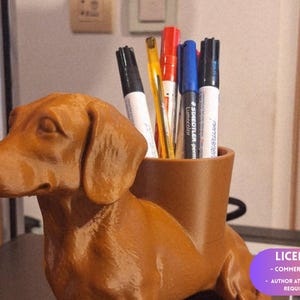 Puede incluir: Un portalápices marrón con forma de perro salchicha con una abertura cilíndrica para bolígrafos y rotuladores. El soporte contiene bolígrafos rojos, azules, negros y amarillos. La figura del perro es de un material liso.