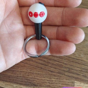 May include: A white keychain with three red circles on a white sphere. The keychain has a black stem and a silver key ring. The keychain is held in a hand.