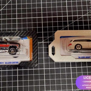 Puede incluir: Dos coches de juguete en embalaje blíster. A la izquierda, un '75 Chevy Blazer Custom negro y rojo en una tarjeta con patrón de fibra de carbono. A la derecha, un '94 Audi Avant RS2 blanco en una tarjeta blanca. Ambos sobre una rejilla negra.