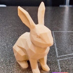 Puede incluir: Una figura de conejo geométrica de color beige claro con facetas angulares y orejas puntiagudas. La escultura presenta un acabado mate y una estructura facetada distintiva que define la forma del animal en estilo low-poly.